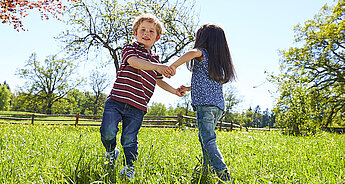 Zwei Kinder, ein Junge und ein Mädchen, spielen auf einer Wiese mit Bäumen im Hintergrund.