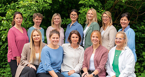 Vier Frauen stehen im Freien vor einer grünen Kulisse, jede trägt unterschiedliche Kleidung und lächelt in die Kamera.
