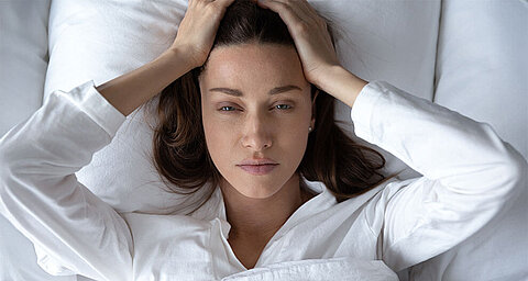 Eine Frau liegt auf einem Bett und hält sich frustriert den Kopf.