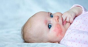 Ein Baby mit blauen Augen, das sich hinlegt und mit der Hand sein Gesicht berührt.