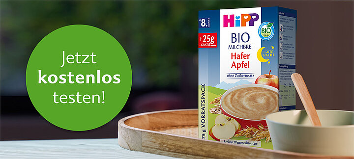 Ein grünes, kreisförmiges Banner mit dem Text "Jetzt kostenlos testen!" ist neben einer Schachtel mit Bio-Babynahrung mit Apfelgeschmack angebracht.