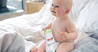 Ein Baby, das auf einem Bett sitzt und eine Flasche mit der Aufschrift "HiPP Milk Lotion Sensitive" in der Hand hält, vor dem Hintergrund eines Zimmers und eines Fensters.