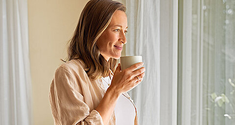 Eine Frau in einem beigen Hemd hält eine weiße Tasse und schaut aus einem Fenster mit durchsichtigen Vorhängen.