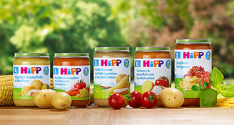 Eine Reihe von Gläschen für Babynahrung mit verschiedenen Geschmacksrichtungen und Zutaten, begleitet von frischen Produkten wie Kartoffeln, Tomaten und Mais.