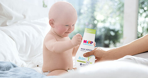 Ein scheinbar gut gelauntes Baby hält ein Fläschchen mit der Aufschrift "HIP BABY SENSITIVE" und der Aufschrift "Milk Lotion".