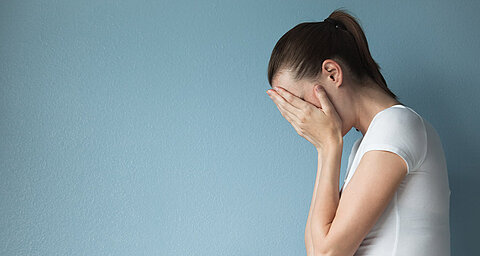 Eine Frau in einem weißen Hemd bedeckt ihr Gesicht mit ihren Händen vor einem blauen Hintergrund.