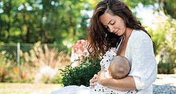 Eine Frau mit langen braunen Haaren, die sanft ein Baby an ihre Brust drückt, im Freien in einer Gartenanlage.
